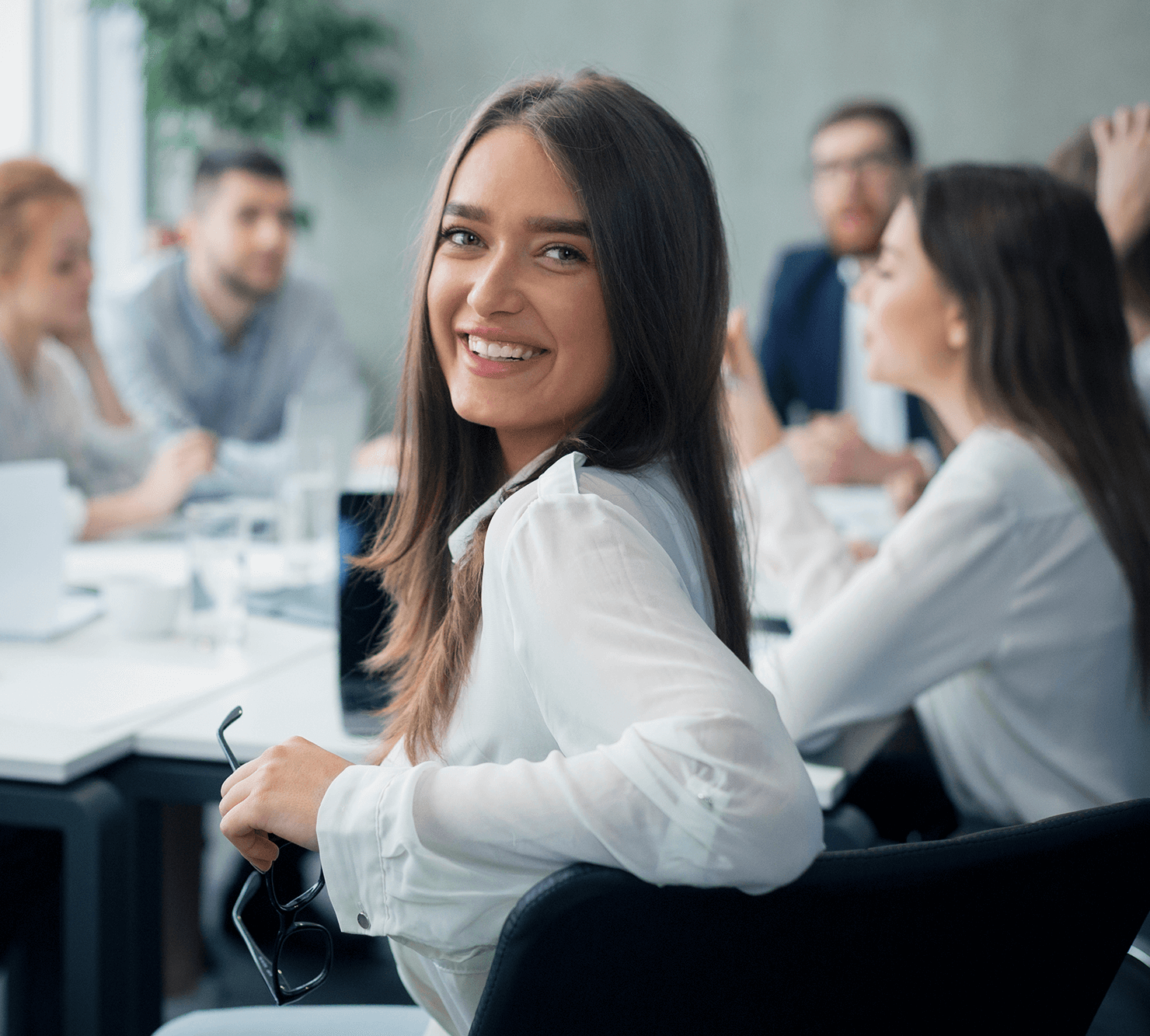 Lächelnde junge Frau in einem Meetingraum, umgeben von Kolleginnen und Kollegen.