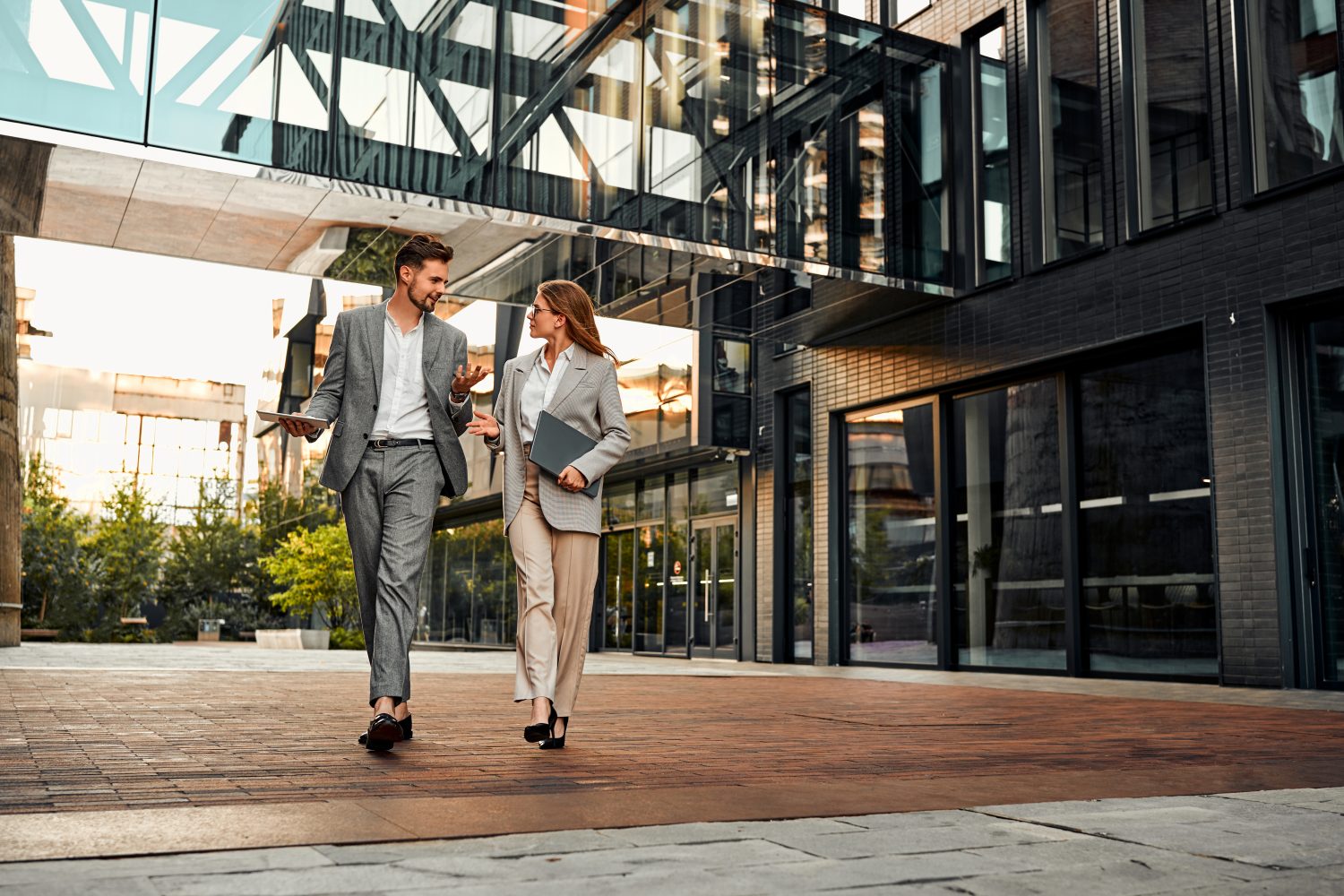 Zwei Business-Mitarbeitende gehen vor einem modernen Bürogebäude entlang und unterhalten sich, beide mit Laptop oder Unterlagen.
