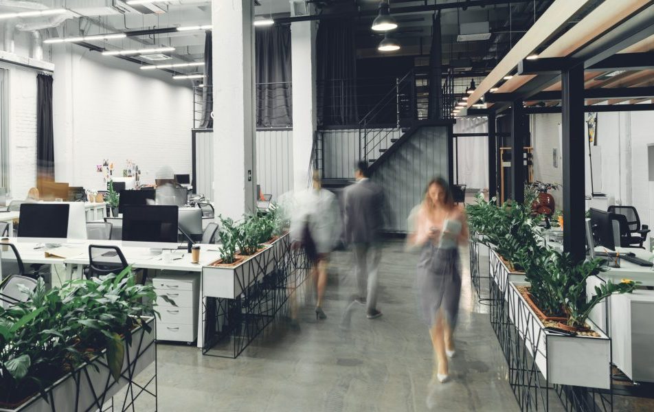 Menschen bewegen sich durch ein modernes Großraumbüro mit Pflanzen und offenen Arbeitsbereichen.