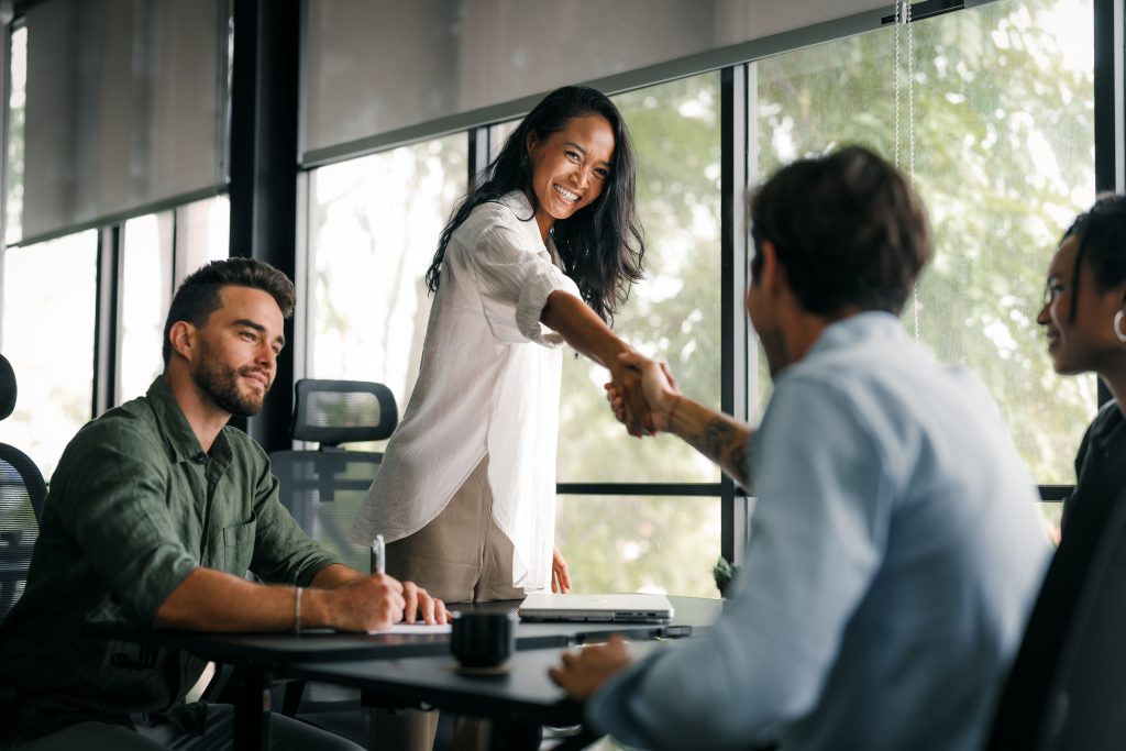 Frau begrüßt einen Kollegen per Handschlag in einem modernen Besprechungsraum, während zwei weitere Personen zusehen.